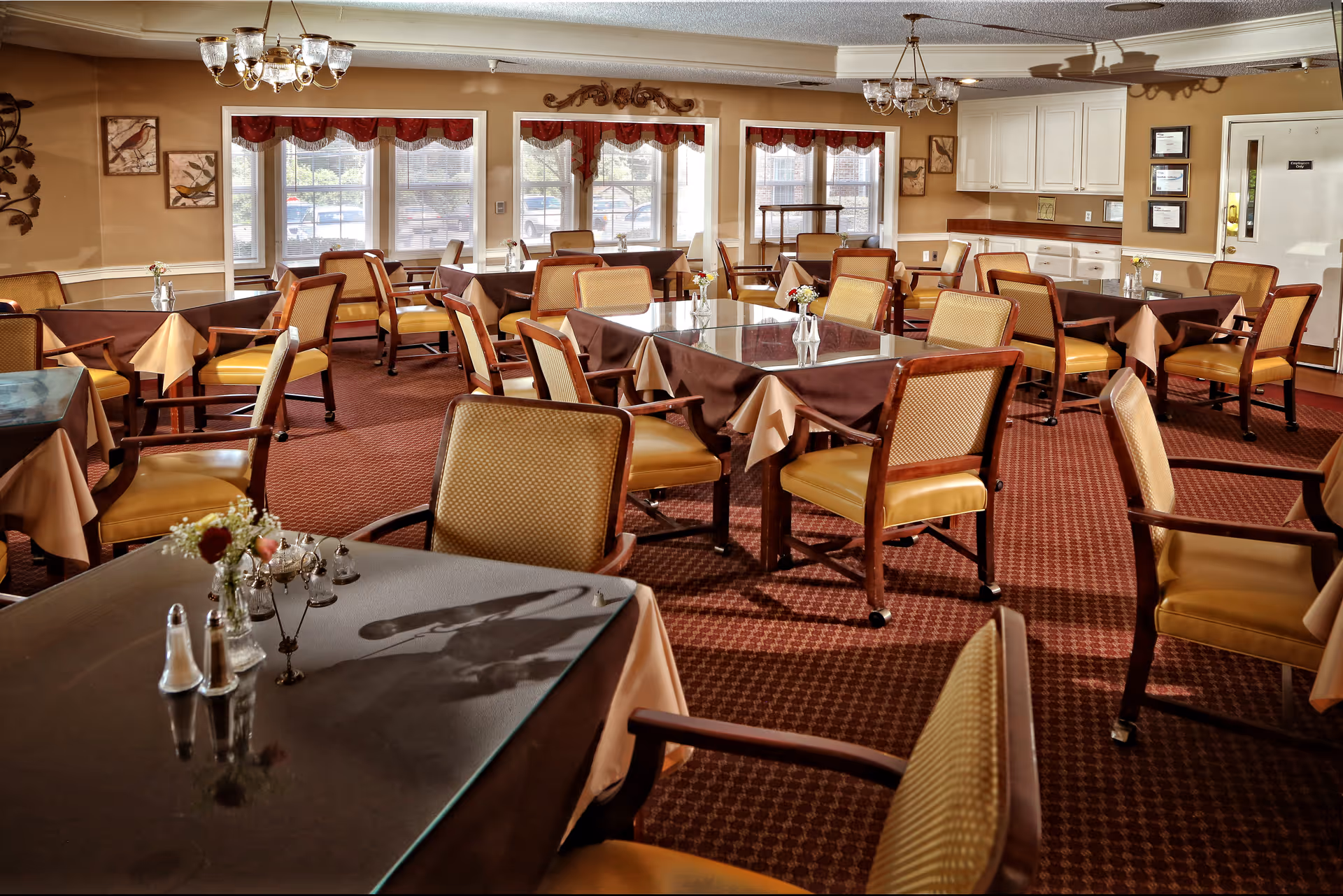 Empty dining room with multiple tables and chairs, table settings, chandeliers, and large windows.