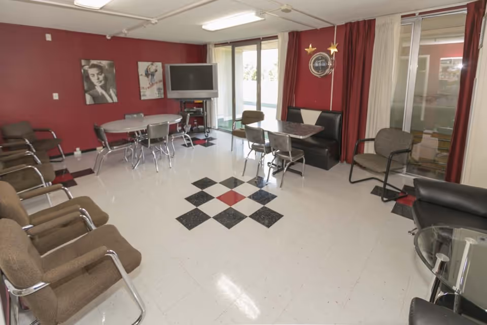 Communal lounge with tables, chairs, a TV and booth seating against red walls and a patterned tile floor.