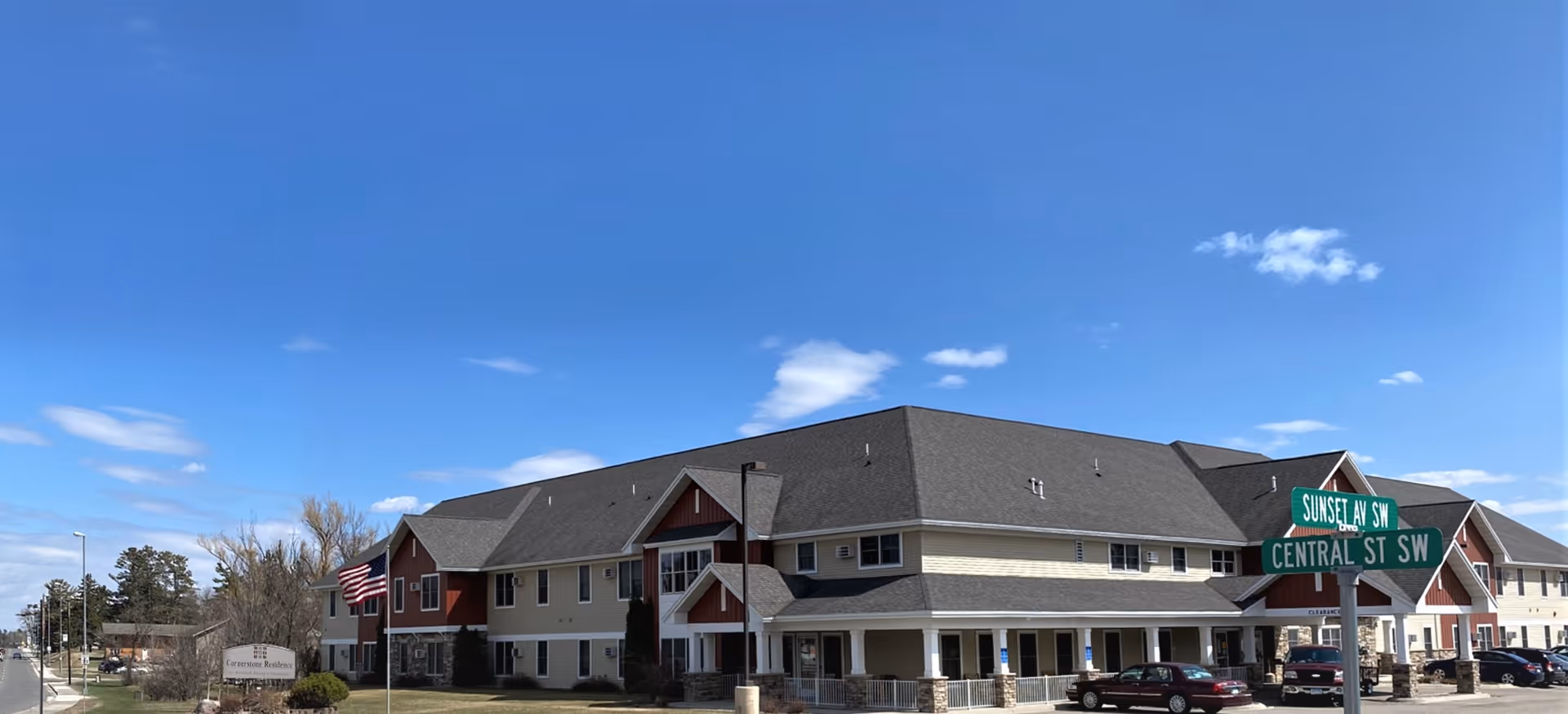 Exterior view of a two-story senior living facility building under a clear blue sky. The building has a combination of beige and red siding with a dark gray roof. An American flag is displayed on a flagpole in front of the building. There is a street sign at the corner showing Sunset Ave SW and Central St SW. Several cars are parked in front of the building.