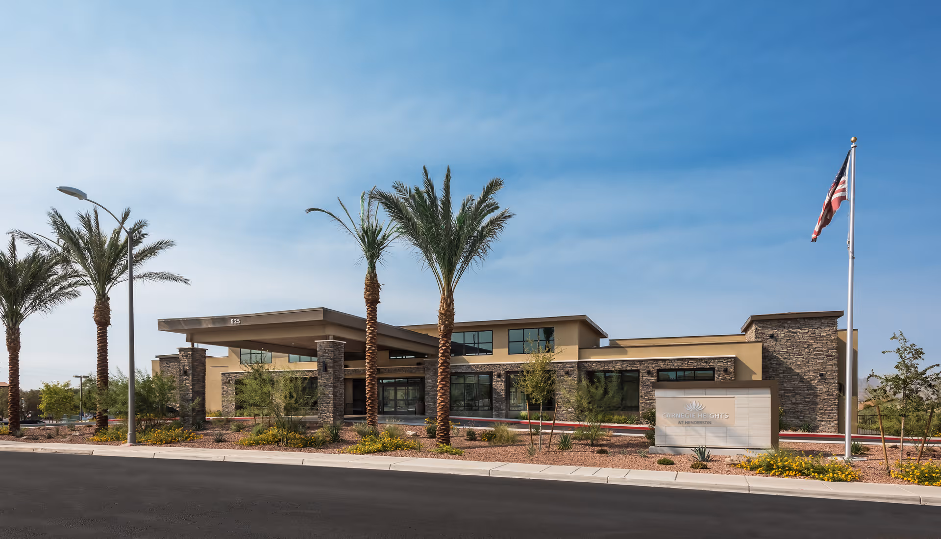 Exterior view of Carnegie Heights at Henderson senior living facility with a modern building featuring stone and beige walls, large windows, palm trees, landscaped desert plants, an American flag on a flagpole, and a clear blue sky.