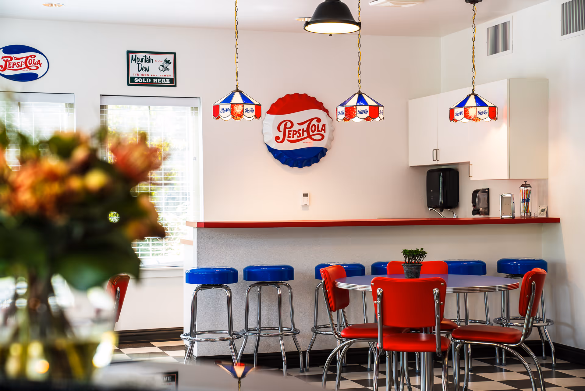 A retro-style dining area with a round table surrounded by red chairs and a counter with blue bar stools. The walls are decorated with vintage Pepsi-Cola signs and a Mountain Dew sign. Three hanging pendant lights with red, white, and blue stained glass shades illuminate the space. A small potted plant is on the table, and a bouquet of flowers is blurred in the foreground.