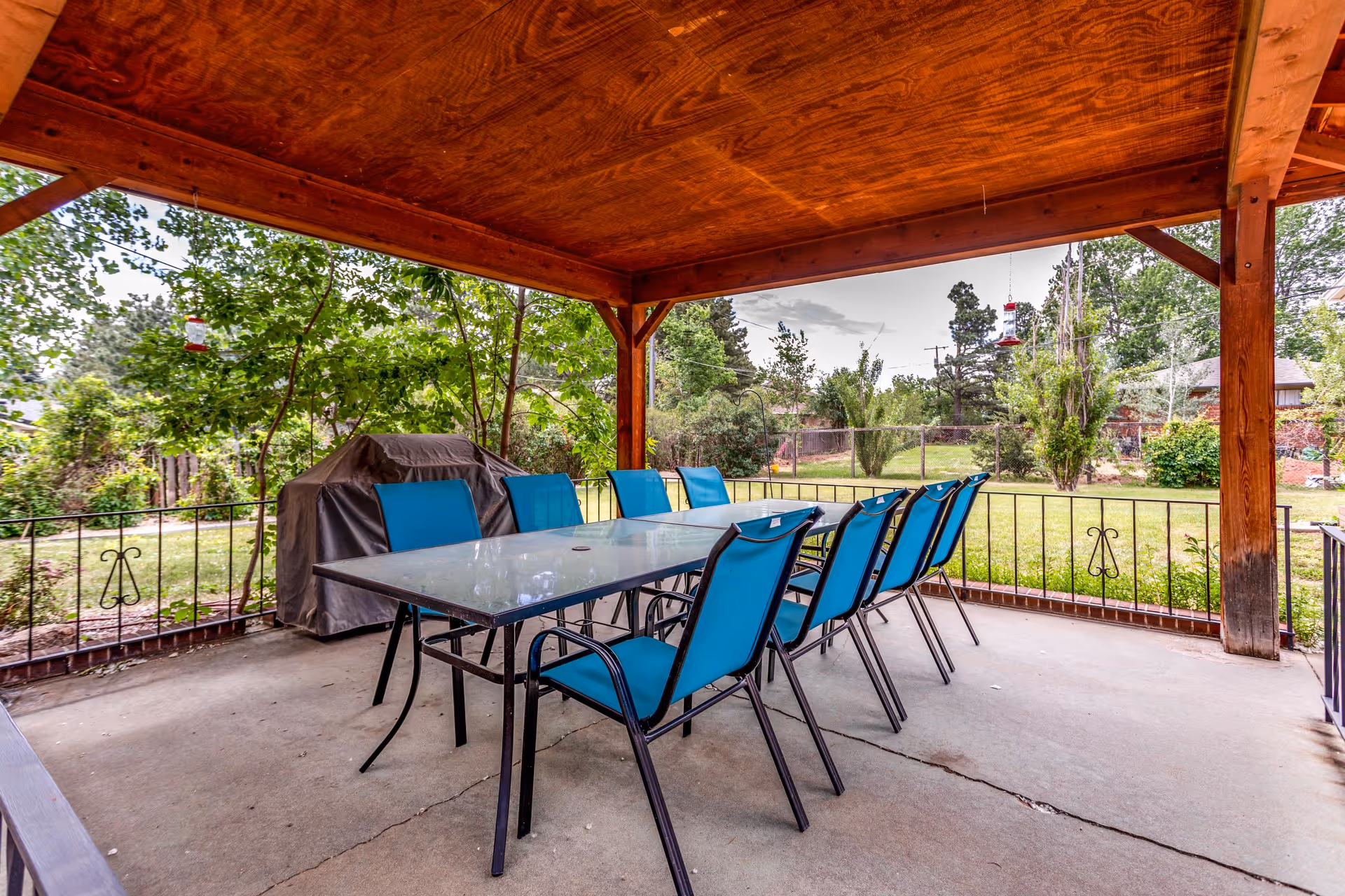 Outdoor covered patio area with a glass-top table surrounded by eight blue chairs. There is a barbecue grill covered with a gray cover in the corner. The patio overlooks a green yard with trees and bushes, enclosed by a metal fence.