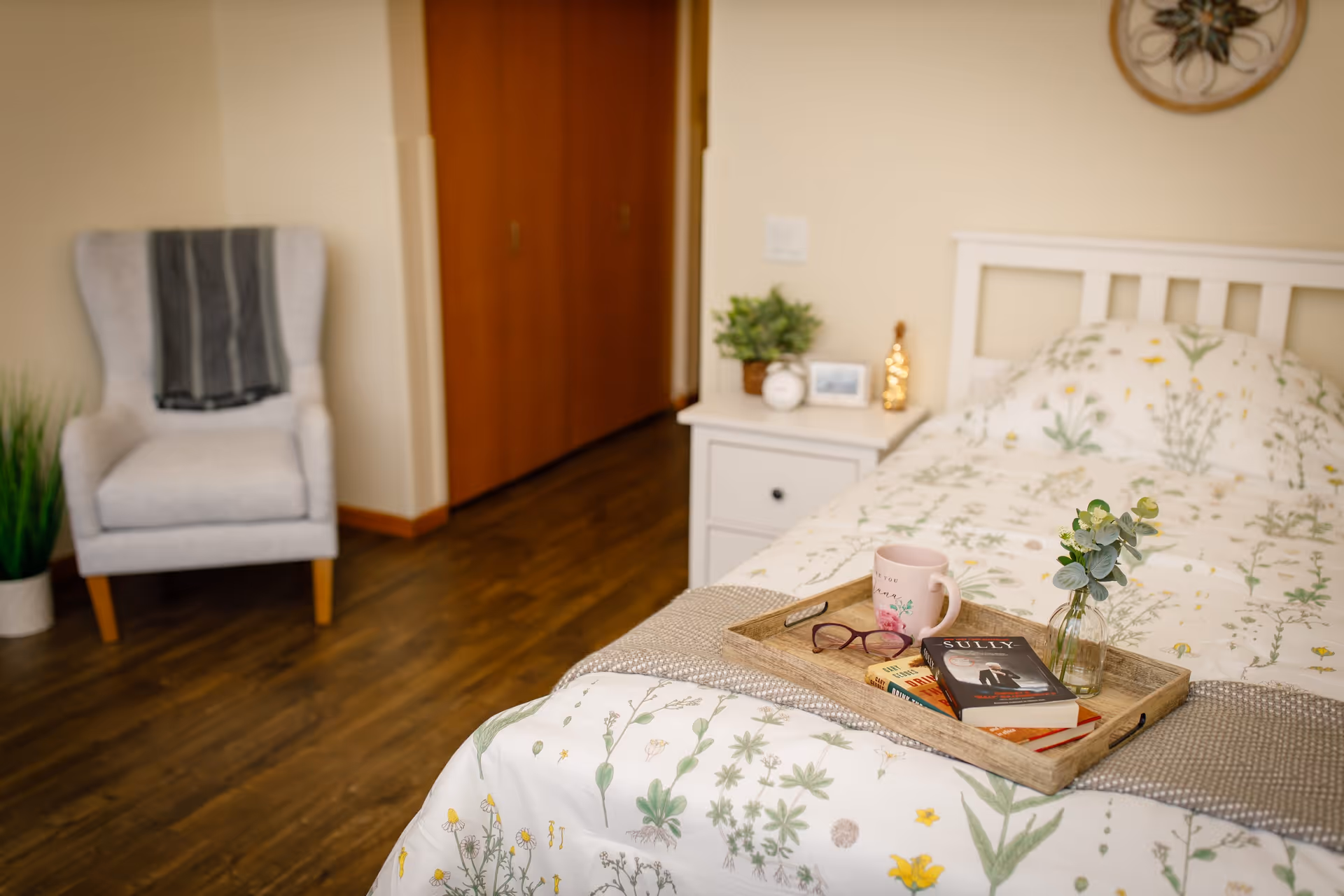 A cozy bedroom with a single bed covered in floral bedding. On the bed is a wooden tray holding a pink mug, a pair of glasses, two books, and a small vase with green foliage. Next to the bed is a white nightstand with a small plant, a framed photo, and a decorative item. In the background, there is a light gray armchair with a dark striped blanket draped over it, a potted plant on the floor, and wooden closet doors.