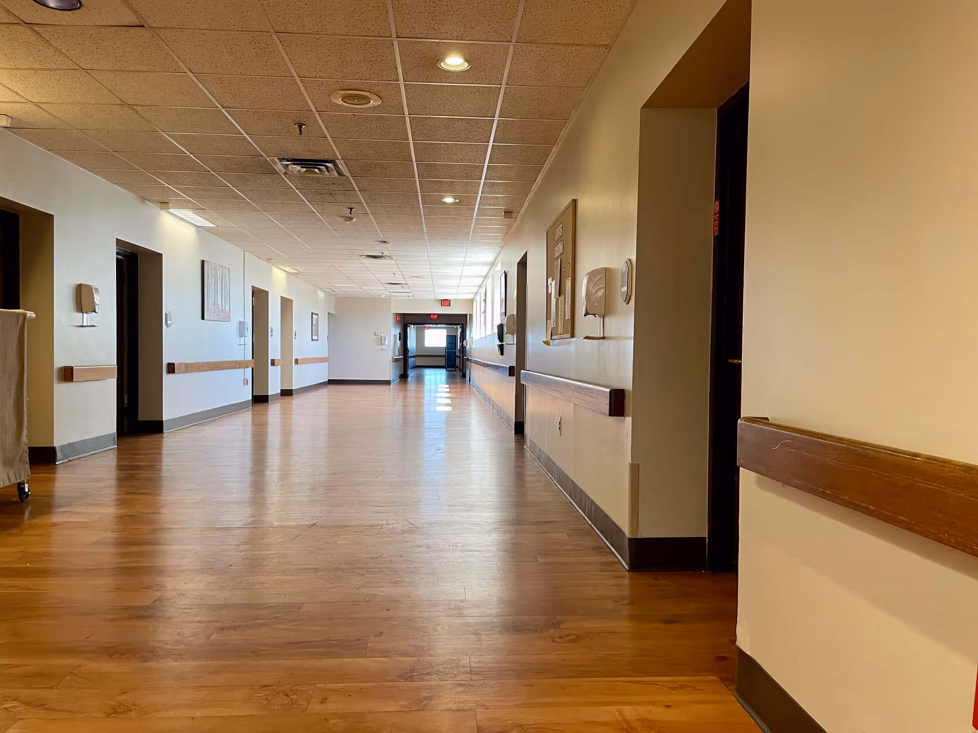 A long, empty hallway inside a healthcare facility with wooden floors, beige walls, and a drop ceiling with recessed lighting. The hallway has several doorways on both sides, handrails along the walls, and a bulletin board on the right wall. Natural light is coming from windows at the far end of the corridor.