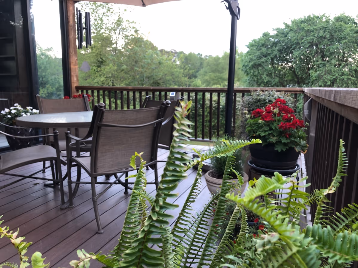 Outdoor patio area with a round glass table and four chairs on a wooden deck. Several potted plants, including ferns and red flowers, are placed around the deck railing. Trees and greenery are visible in the background.