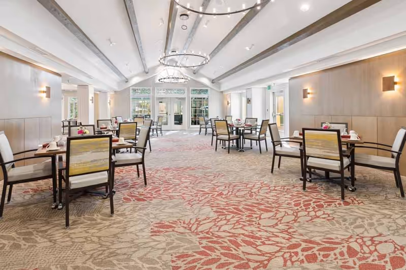 A spacious dining room with multiple tables and chairs arranged neatly. The room features a patterned carpet with red and beige floral designs, light-colored walls with wooden paneling, and large windows at the far end letting in natural light. Modern chandeliers hang from the ceiling, and each table is set with cups and small flower arrangements.