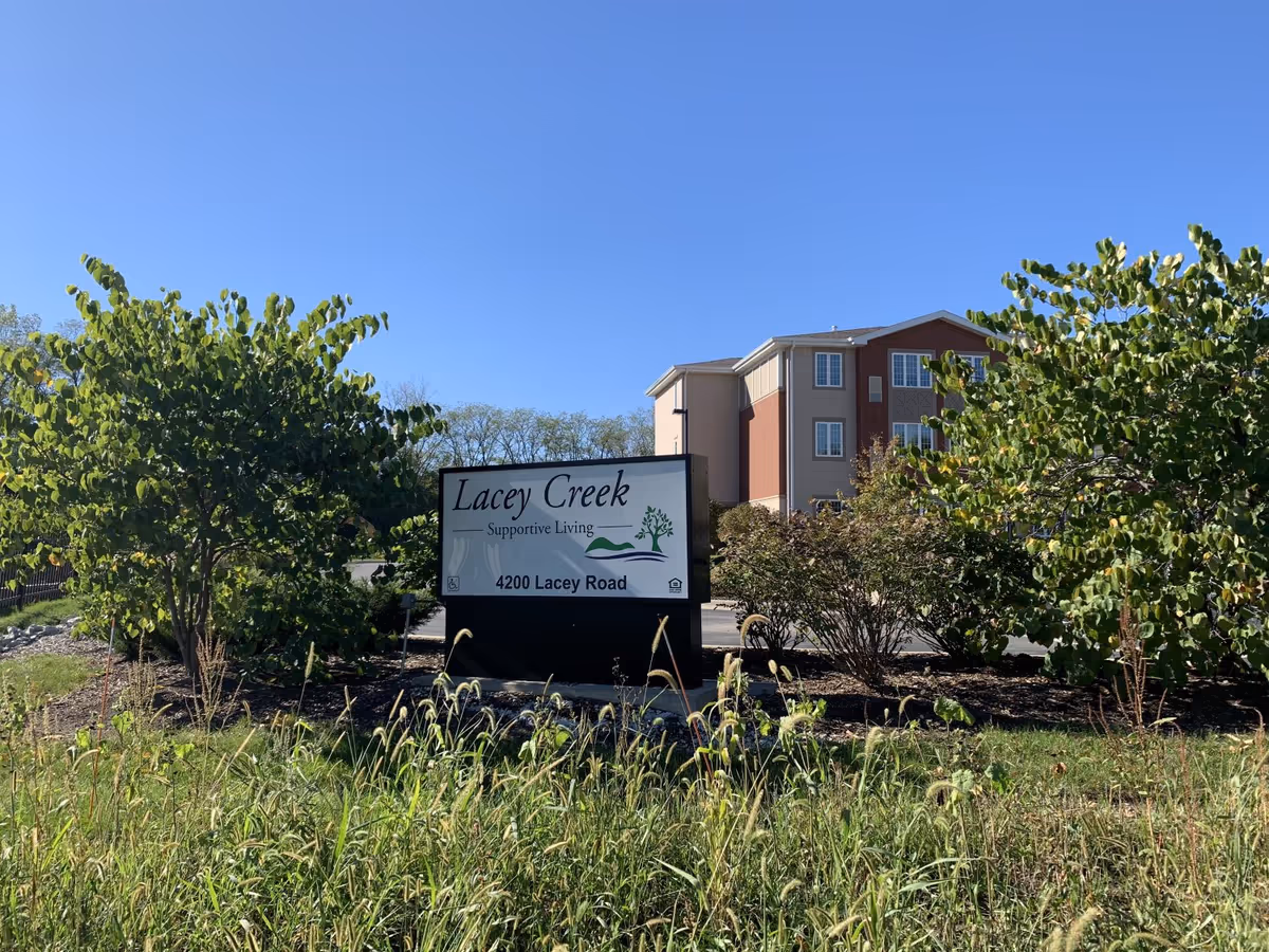 Outdoor view of the entrance sign for Lacey Creek Supportive Living located at 4200 Lacey Road, surrounded by greenery and bushes with a multi-story building in the background under a clear blue sky.