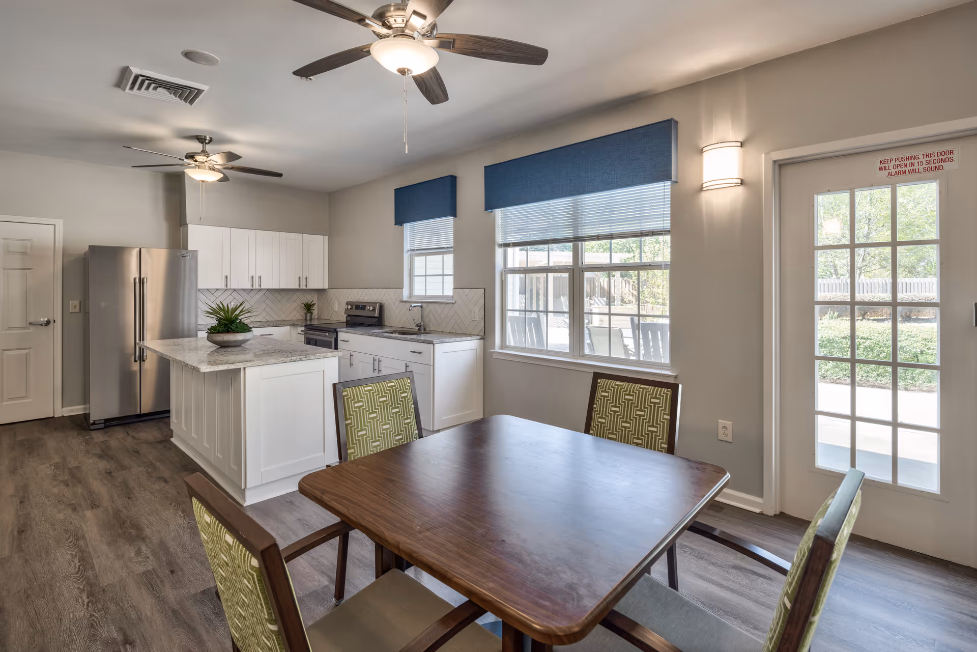 A bright kitchen and dining area with white cabinets, a stainless steel refrigerator, an island with a plant centerpiece, and a wooden dining table with four green patterned chairs. Two ceiling fans with lights are mounted on the ceiling. Large windows with blue valances allow natural light to fill the room, and a glass door leads outside to a patio area with rocking chairs.