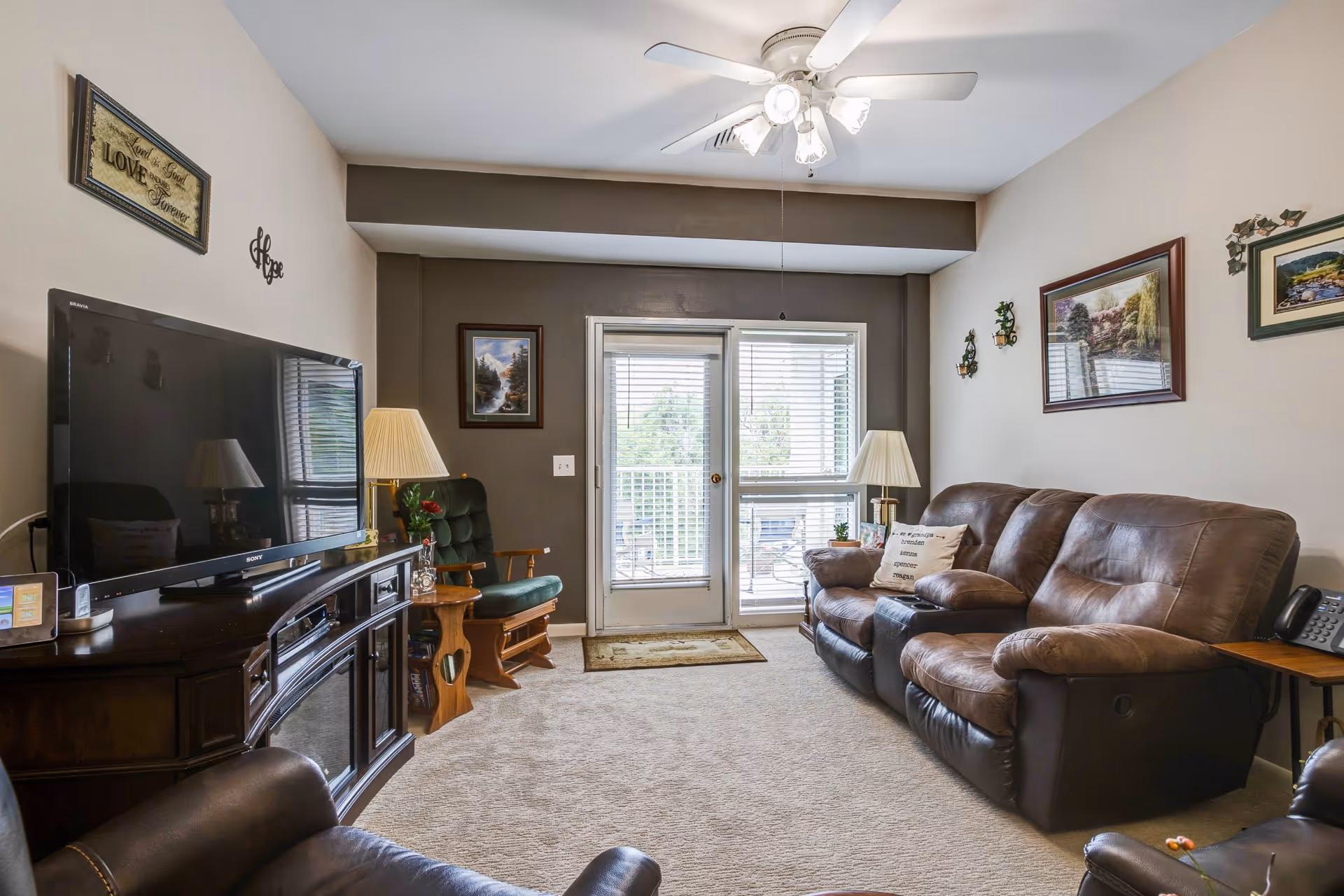 A cozy living room with a brown leather sofa, a green cushioned rocking chair, a dark wooden TV stand with a flat-screen TV, two table lamps, framed landscape paintings on the walls, and a ceiling fan with lights. There is a glass door and window with blinds leading to an outdoor area.