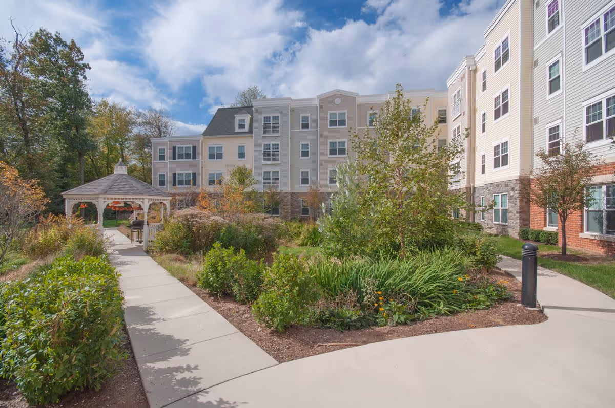 Outdoor garden area with a concrete walkway leading to a white gazebo surrounded by greenery and bushes, with a multi-story senior apartment building in the background under a partly cloudy sky.