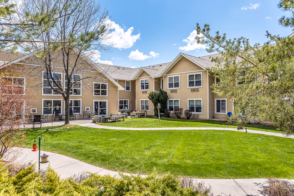 Sunny courtyard with a green lawn, paved paths, patio seating, and a two-story beige senior living building under a blue sky.
