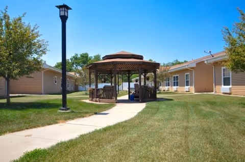 Outdoor area of Windsor Manor featuring a wooden gazebo with benches, surrounded by green grass and trees, with beige single-story buildings on either side under a clear blue sky.