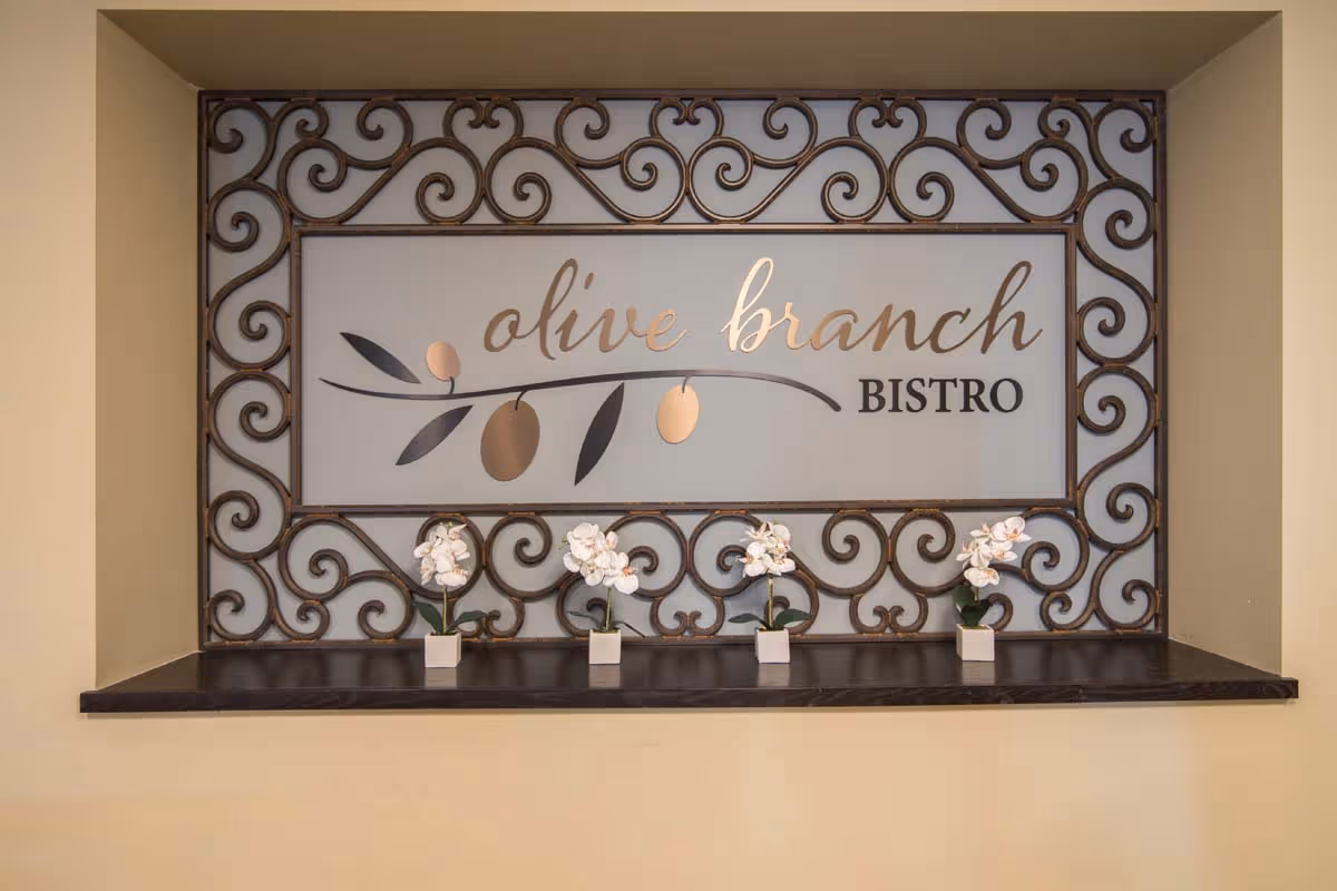 Decorative 'olive branch Bistro' sign framed with wrought-iron scrollwork above a shelf holding four small potted white orchids.