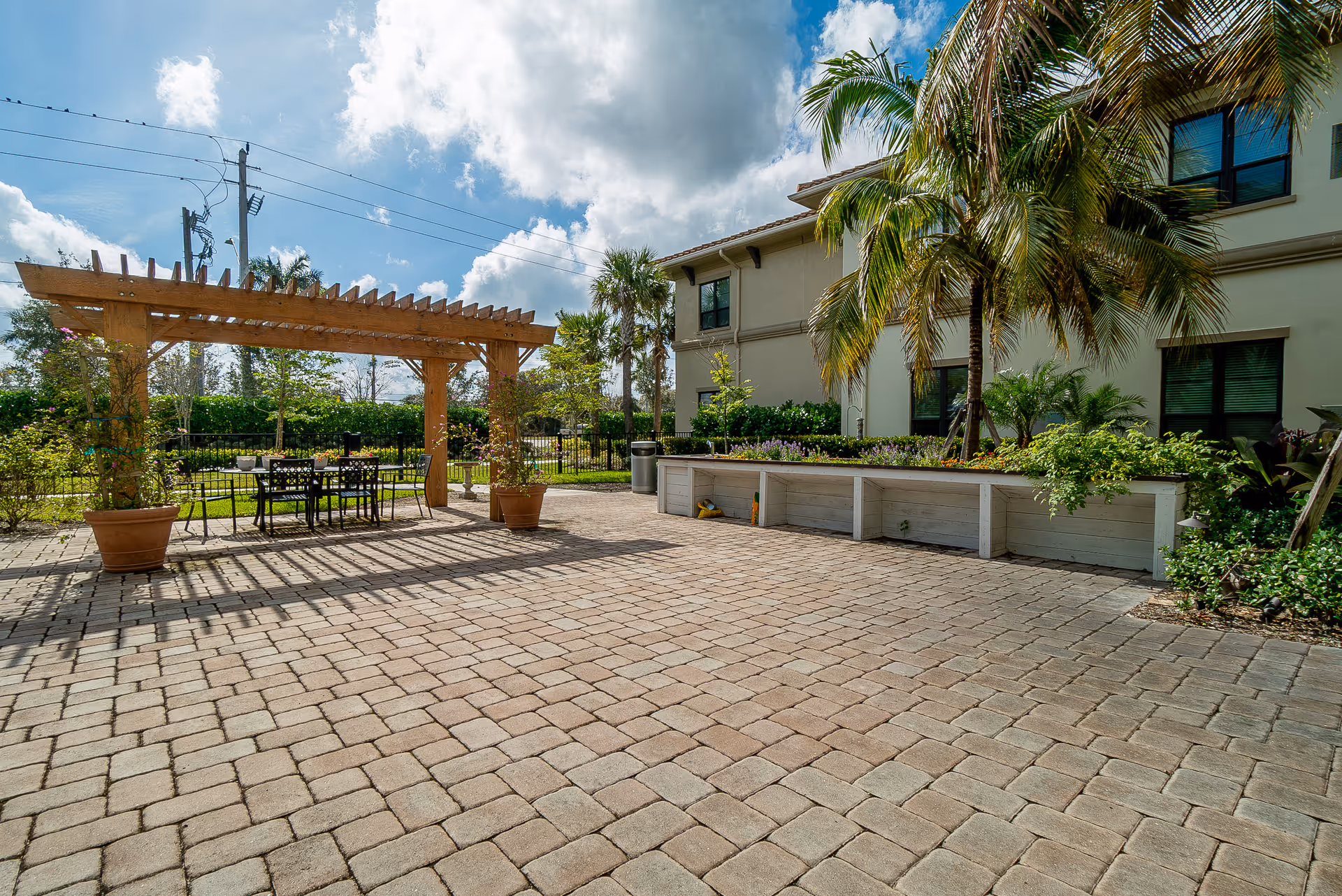 Outdoor patio area with a wooden pergola, a table and chairs underneath, surrounded by potted plants and greenery. The patio is paved with stone tiles and adjacent to a building with windows and palm trees.