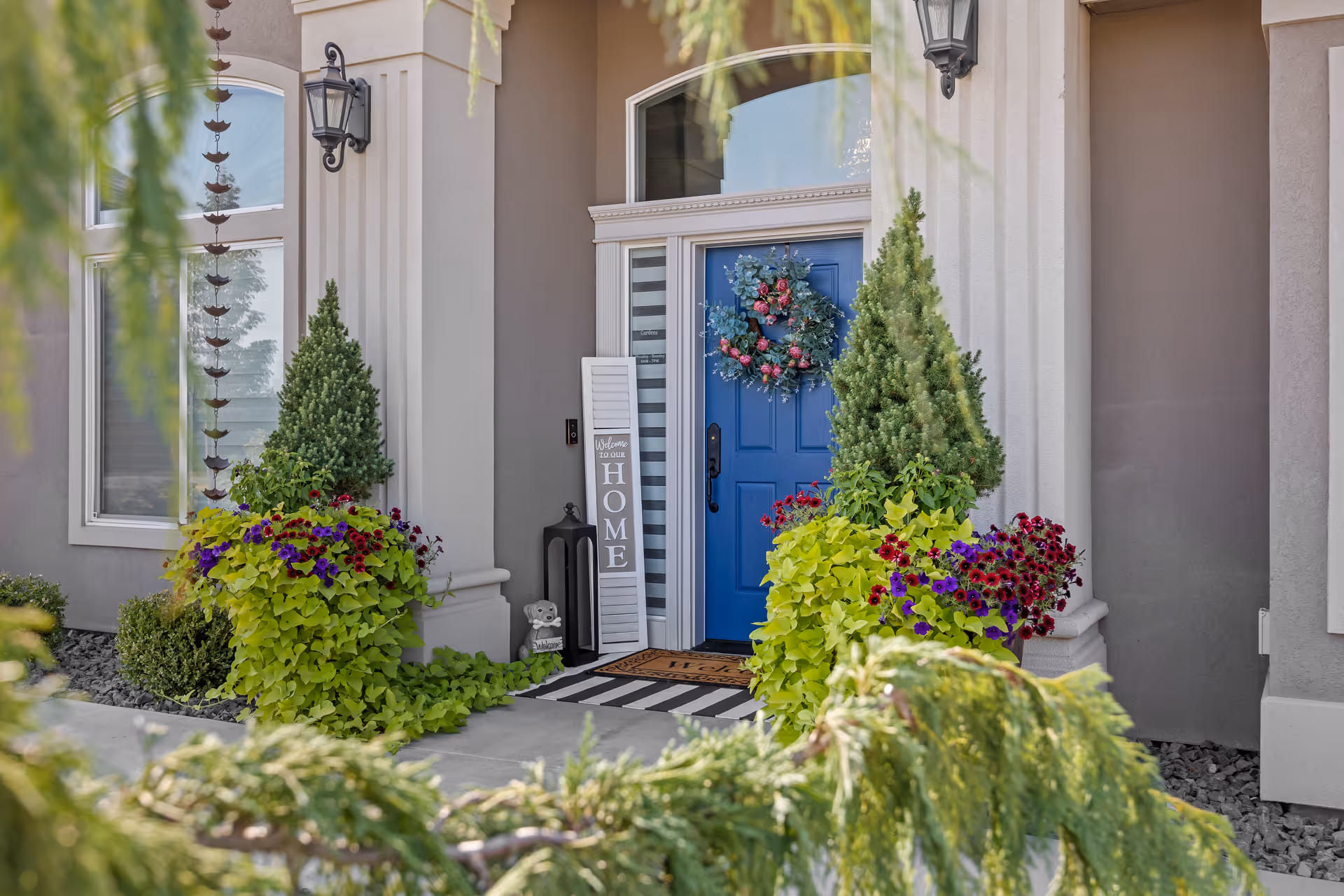 Front entrance of a house with a blue door decorated with a wreath, potted plants, and a welcome sign.