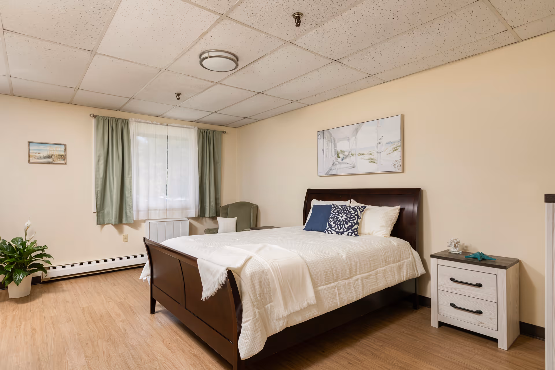 Furnished bedroom with a wooden bed, white bedding, a nightstand, armchair, window with green curtains, and wall art.
