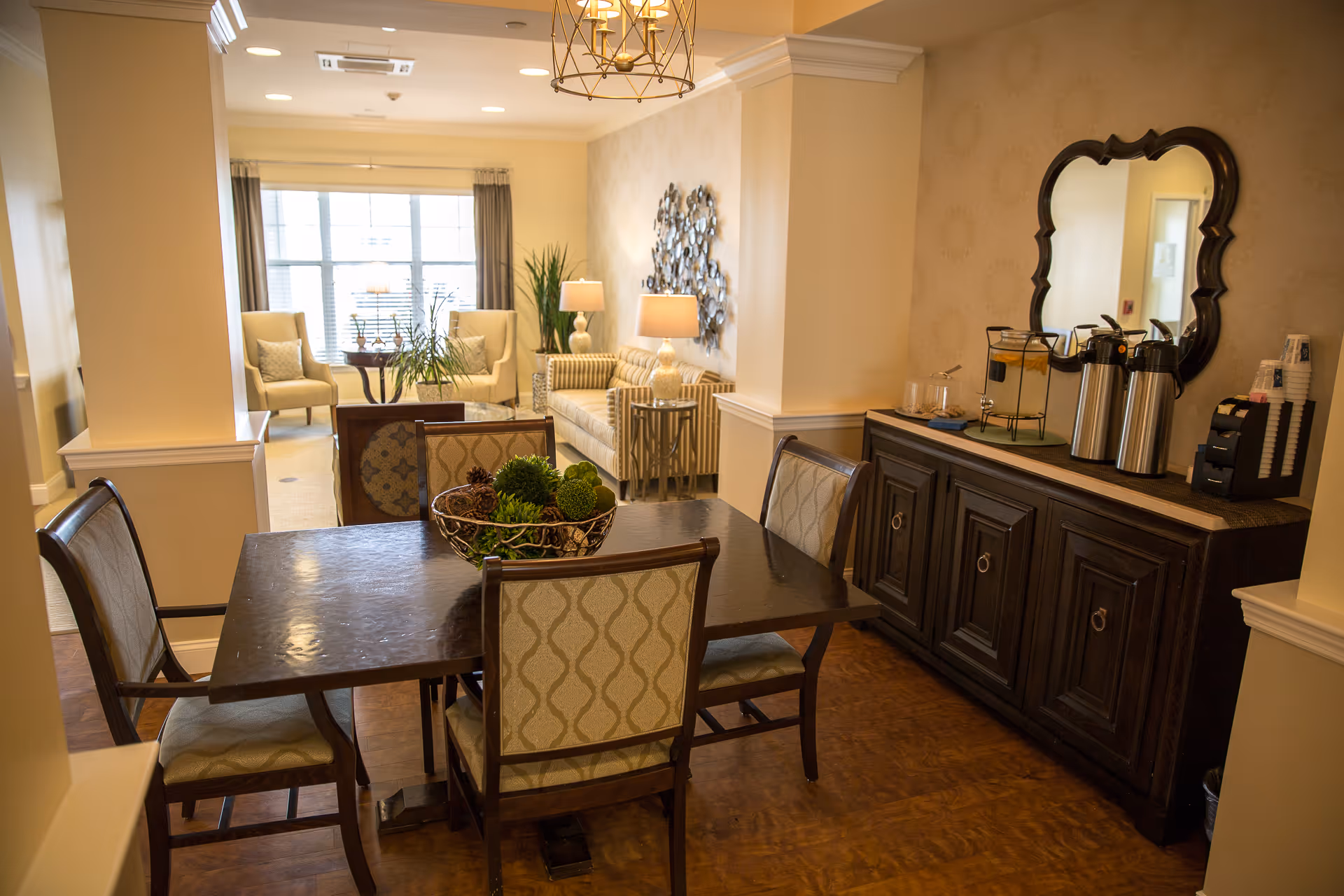 A cozy and well-lit interior space featuring a dark wooden dining table with four upholstered chairs. On the table is a decorative bowl with green and brown natural elements. To the right, there is a dark wooden sideboard with a large ornate mirror above it, holding coffee dispensers, cups, and other beverage supplies. In the background, a sitting area with a striped sofa, two armchairs, side tables with lamps, plants, and a large window with curtains is visible.