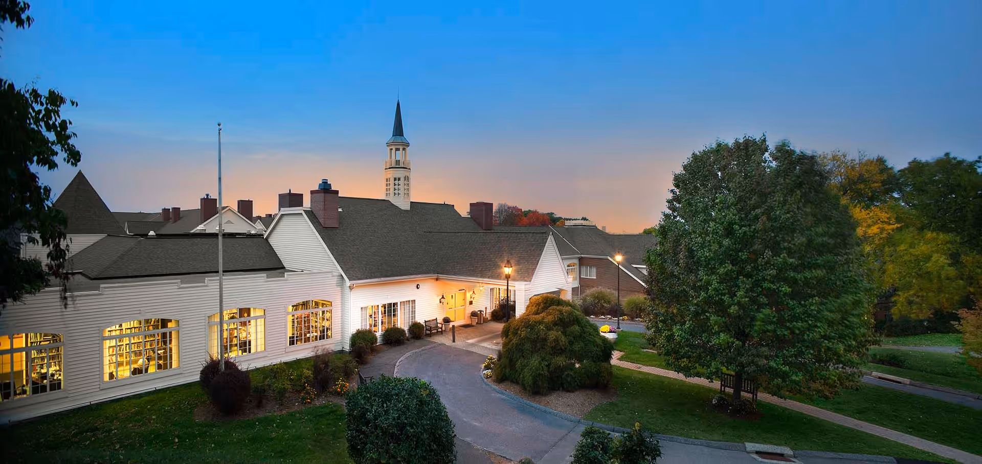 Exterior view of a senior living facility named Avery Heights at dusk, showing a large white building with multiple windows lit from inside, a tall steeple, surrounding greenery including trees and bushes, and a curved driveway leading to the entrance.