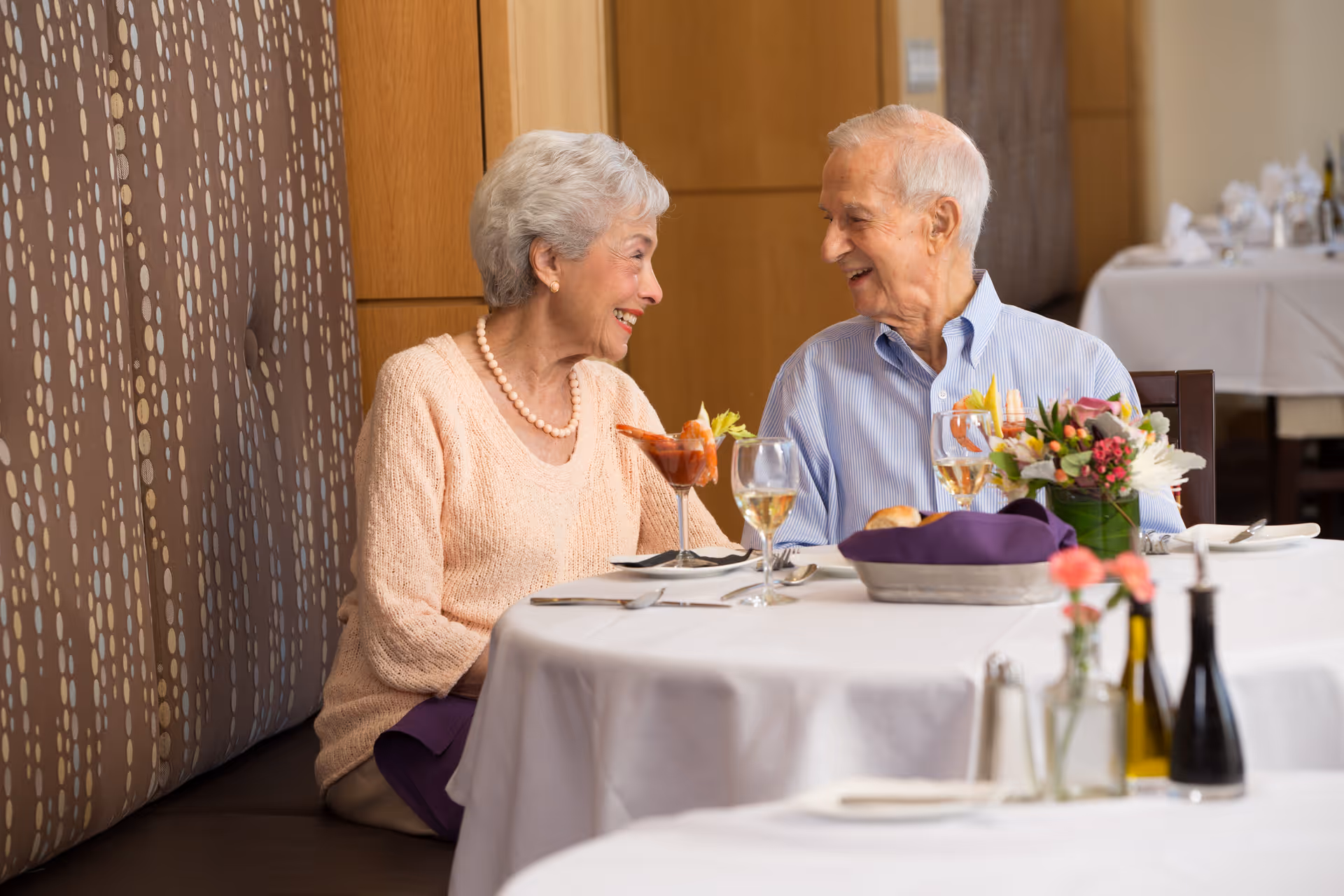 An elderly woman and man sitting at a dining table in a restaurant or dining room, smiling and engaging in conversation. The table is set with glasses of wine, a cocktail, a bread basket, and a small floral arrangement. The background shows a cushioned bench and wooden paneling.