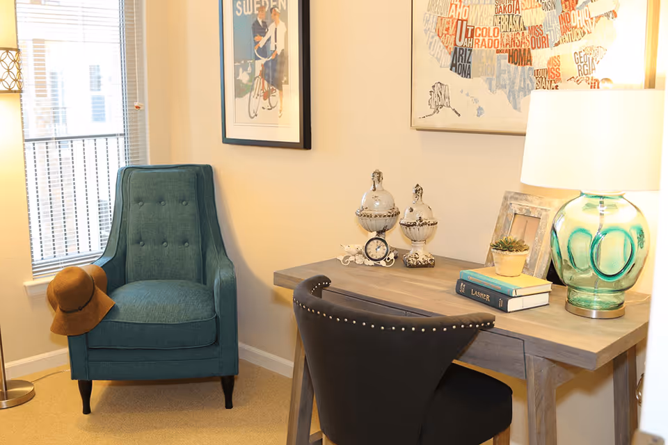 A cozy corner of a room featuring a teal upholstered armchair with a brown hat resting on one arm, next to a window with blinds. Adjacent to the chair is a wooden desk with a black studded chair. On the desk are decorative items including two ornate jars, a small clock, a framed picture, a small potted plant, two books, and a green glass table lamp. The walls are adorned with framed artwork, one depicting a map of the United States and another showing a person riding a bicycle.