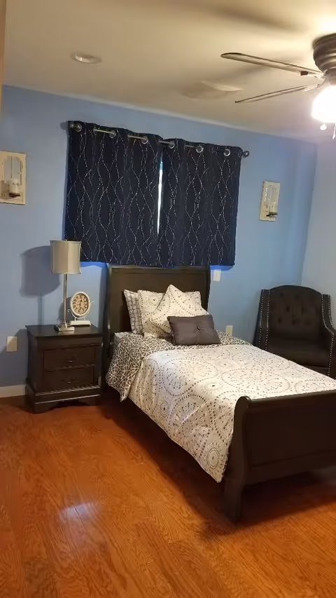 A bedroom with light blue walls and wooden flooring featuring a single dark wooden bed with patterned white and gray bedding and multiple pillows. Next to the bed is a dark wooden nightstand with a lamp and a clock. There is a dark upholstered armchair in the corner and a ceiling fan with a light above. The window has dark blue curtains with a wavy pattern.