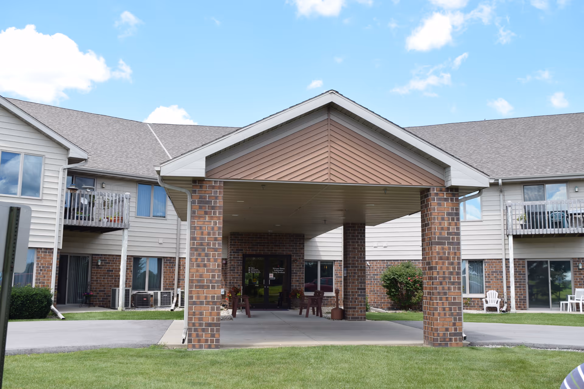 Front entrance of a two-story senior living facility with a covered drop-off area supported by brick pillars. The building has a combination of brick and light-colored siding, with balconies and windows visible. There is a well-maintained lawn and a clear blue sky with some clouds.