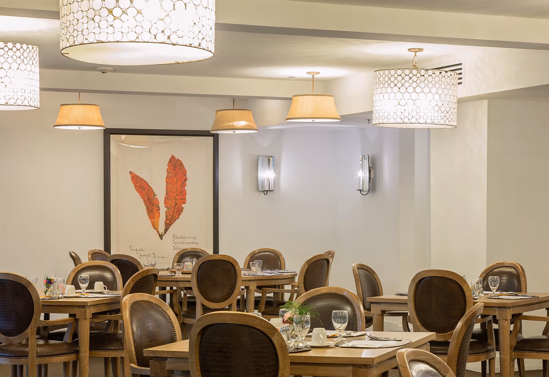 A dining room with multiple wooden tables and chairs arranged neatly. Each table is set with glassware, cups, and small flower arrangements. The room is softly lit by several ceiling lamps with decorative shades, and there is a framed artwork featuring two large red leaves on the wall.