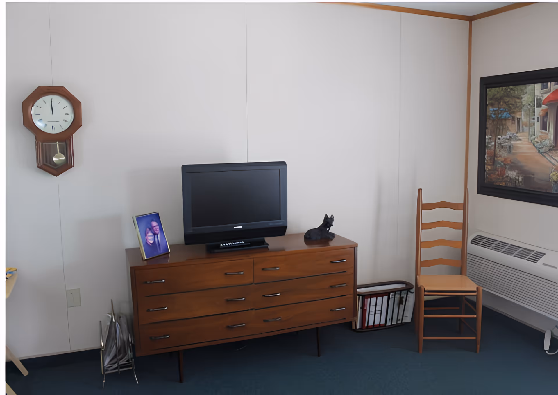A small room with a wooden dresser holding a flat-screen TV, a framed photo, and a small dog figurine. A wooden chair is placed next to the dresser near a window with a painting above a wall-mounted heater. A wall clock hangs on the left wall.