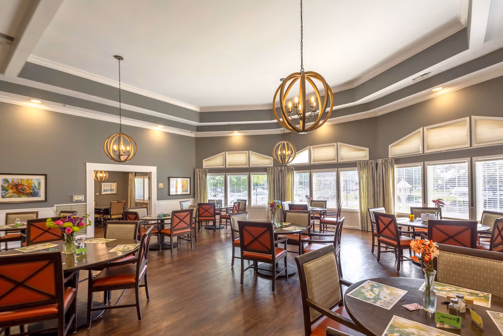 Spacious dining room with round tables and chairs, decorative pendant lights, large windows, and floral centerpieces.