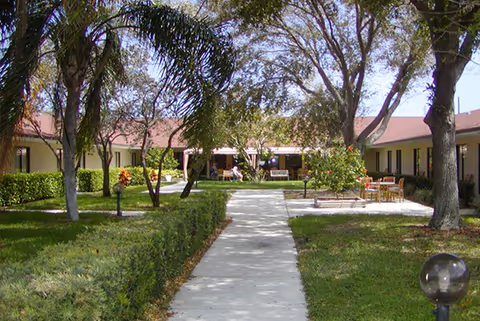 Outdoor courtyard area with a concrete walkway leading through green grass and trees, surrounded by single-story buildings with red roofs. There are benches and tables with chairs placed on the grass near the buildings.