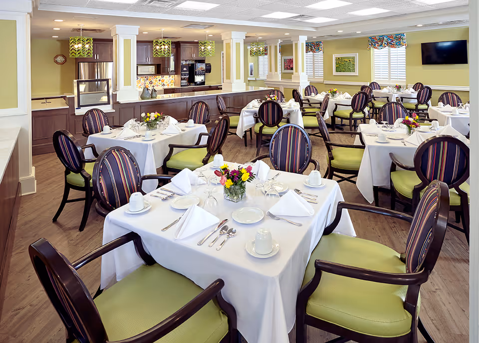A bright and elegant dining room with multiple tables covered in white tablecloths, each set with plates, cups, silverware, and napkins. The chairs have dark wooden frames with green cushions and striped backs. The room features wooden flooring, green walls with white trim, decorative pendant lights, and a kitchen area with dark cabinetry and stainless steel appliances in the background.