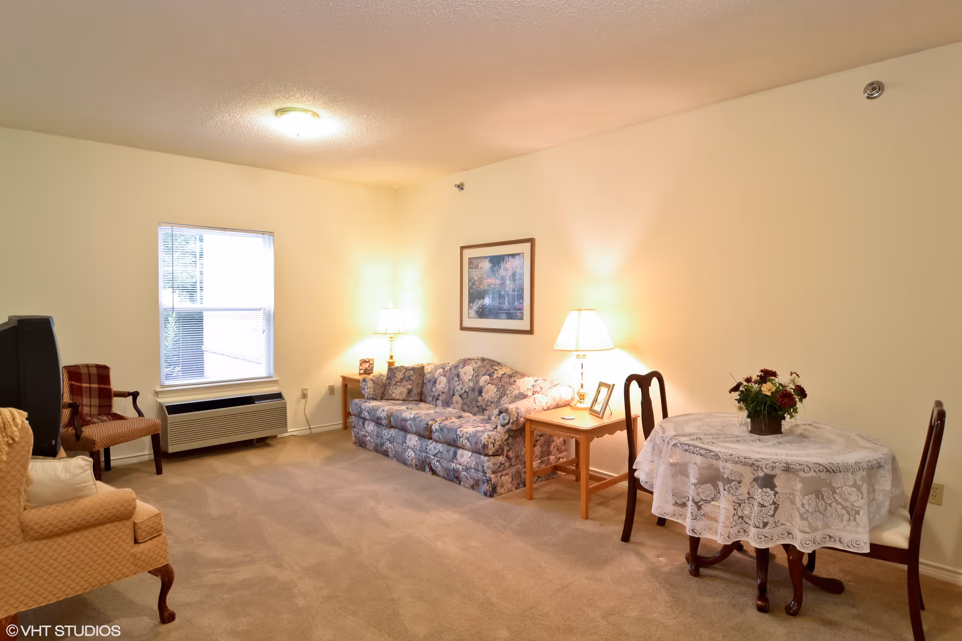 A cozy living room with beige carpet and cream-colored walls. The room features a floral patterned sofa with two side tables, each holding a lamp. There is a framed picture hanging above the sofa. To the right, there is a round table covered with a white lace tablecloth and decorated with a flower arrangement, surrounded by two wooden chairs. On the left side, there is a cushioned armchair and a TV on a stand near a window with blinds. The room is softly lit by ceiling and table lamps.