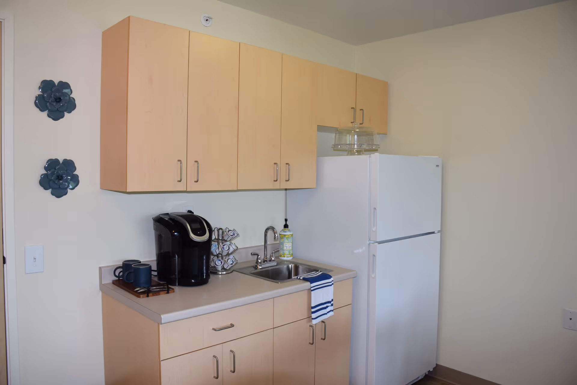 Small kitchenette with light wood cabinets, a white refrigerator, a sink with a striped towel hanging over the edge, a black coffee maker, and a coffee pod holder. Two decorative blue flower wall hangings are on the left wall.