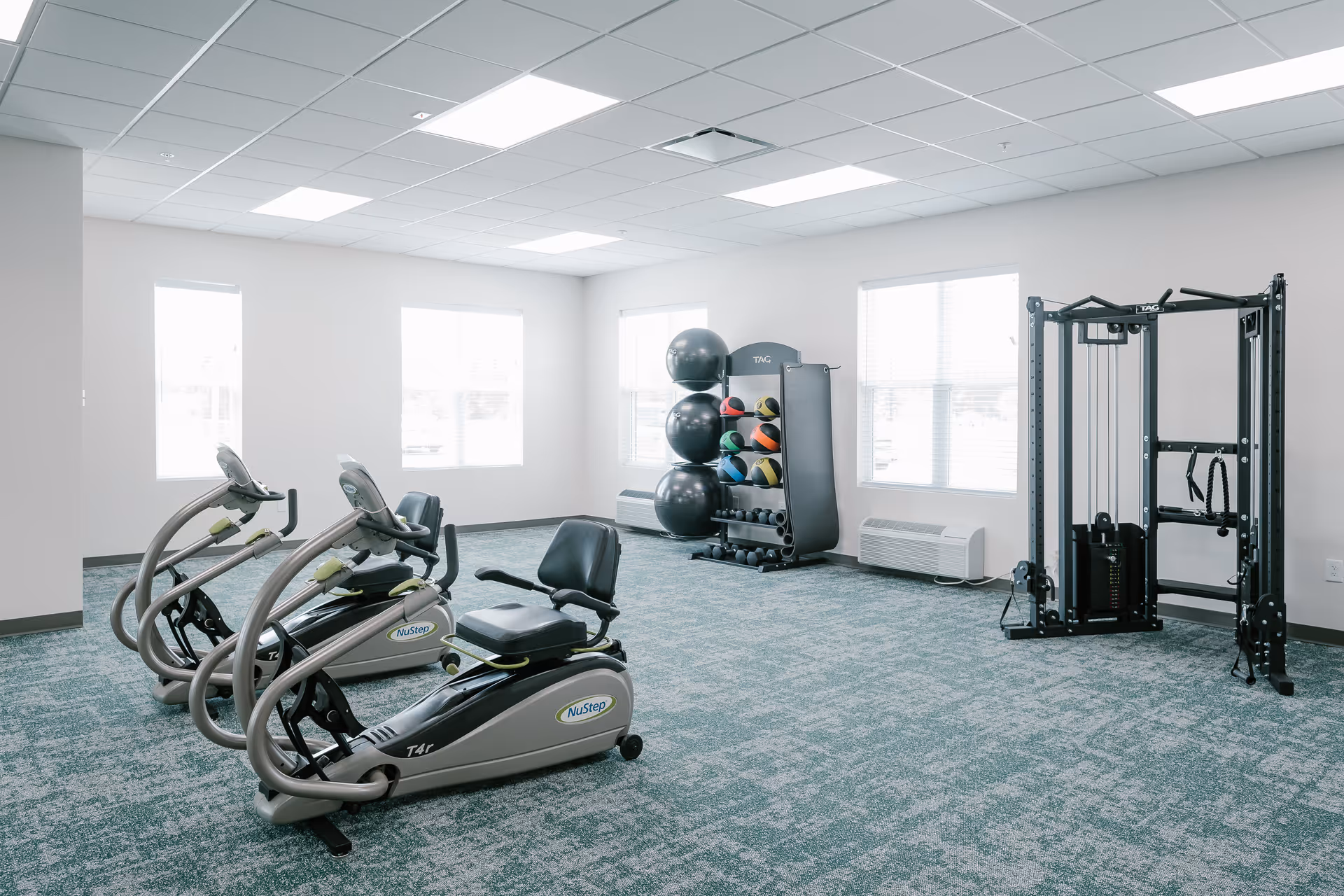 A bright exercise room with three NuStep recumbent cross trainers, a rack holding large exercise balls and colorful medicine balls, and a multi-functional cable machine. The room has large windows letting in natural light and a patterned carpet floor.