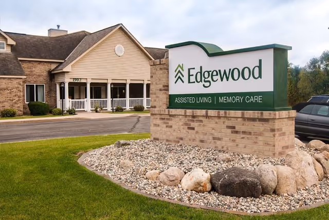 Exterior view of Edgewood Alexandria senior living facility showing a brick building with a covered entrance and a large sign in the foreground that reads 'Edgewood Assisted Living | Memory Care'. The sign is surrounded by rocks and landscaping with green grass.