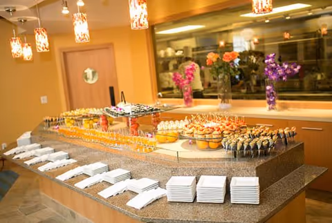 A buffet table in a dining area set with various appetizers and desserts in small glasses and plates, with stacks of white napkins and plates arranged neatly along the edge. The room has warm lighting with hanging pendant lights and floral arrangements in the background.