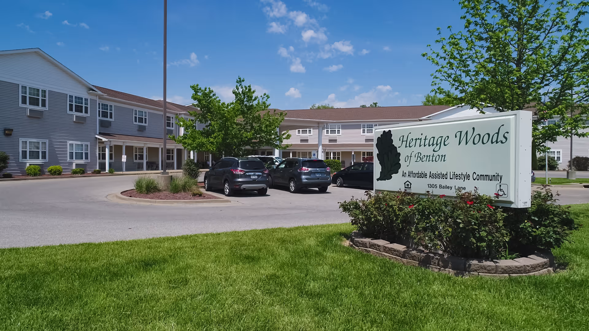 Exterior view of Heritage Woods of Benton, an affordable assisted lifestyle community, showing a two-story building with multiple windows, a parking lot with several cars, green grass, trees, and a clear blue sky.