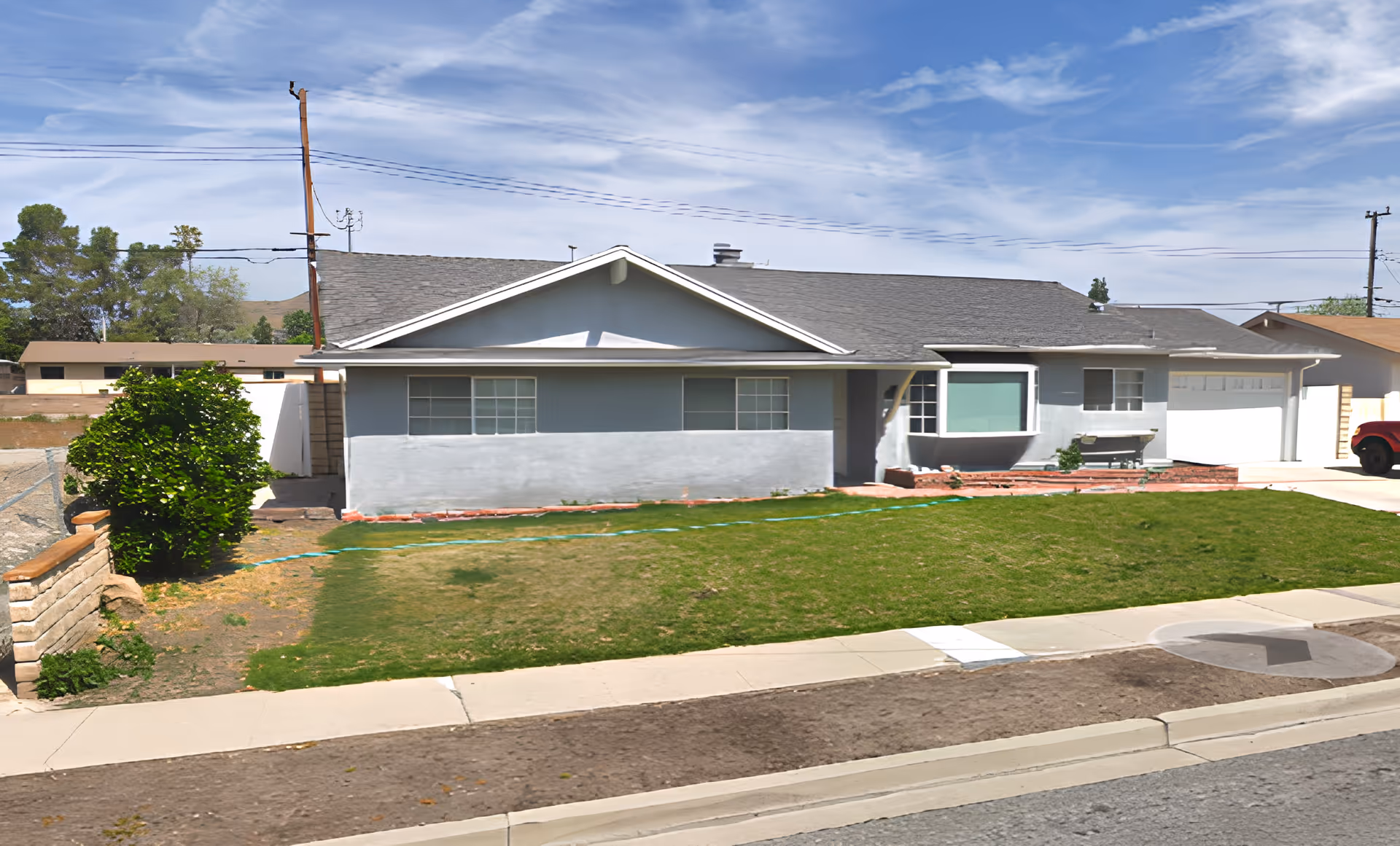 Single-story residential building with a gray exterior and a sloped roof, featuring a front lawn with patchy grass, a small bush on the left side, a driveway leading to a garage on the right, and a clear blue sky with some clouds above.