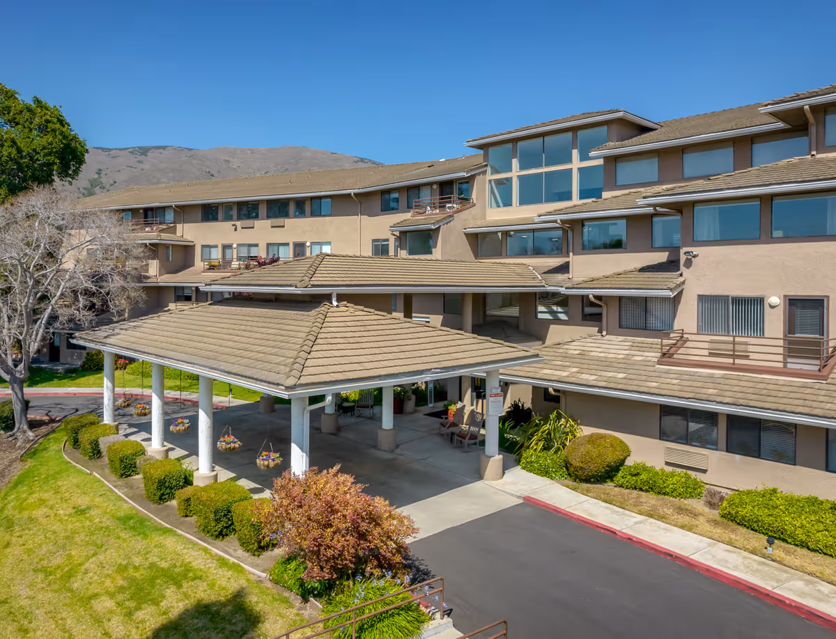 Front entrance of a multi-story beige senior living building with a covered porte-cochere, landscaped shrubs, and hills in the background.
