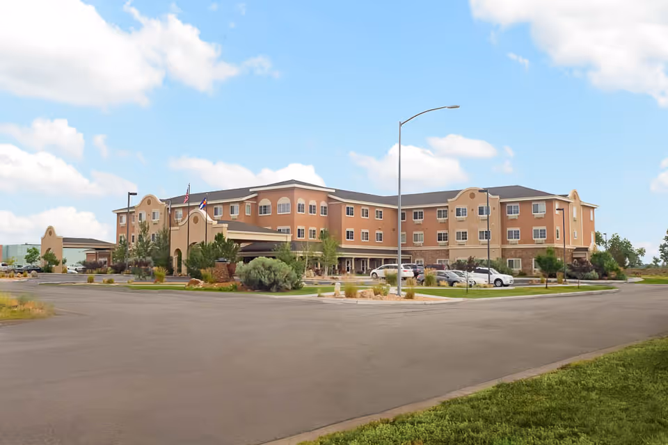 Exterior view of a three-story senior living facility building with a beige and brown facade, surrounded by a parking lot with several cars and landscaped greenery under a partly cloudy sky.