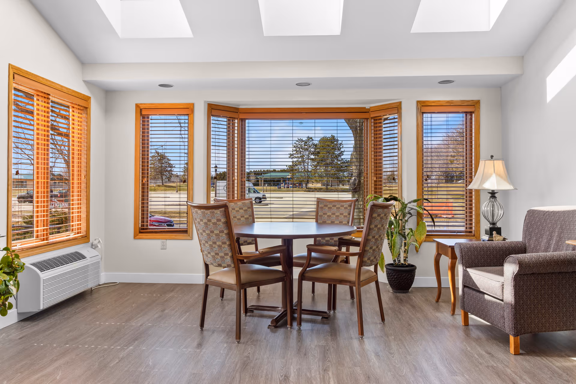 A bright room with large windows featuring wooden blinds, a round table with four cushioned chairs, a potted plant, a side table with a lamp, and an upholstered armchair. The room has light-colored walls and wood flooring, with skylights allowing natural light to fill the space.