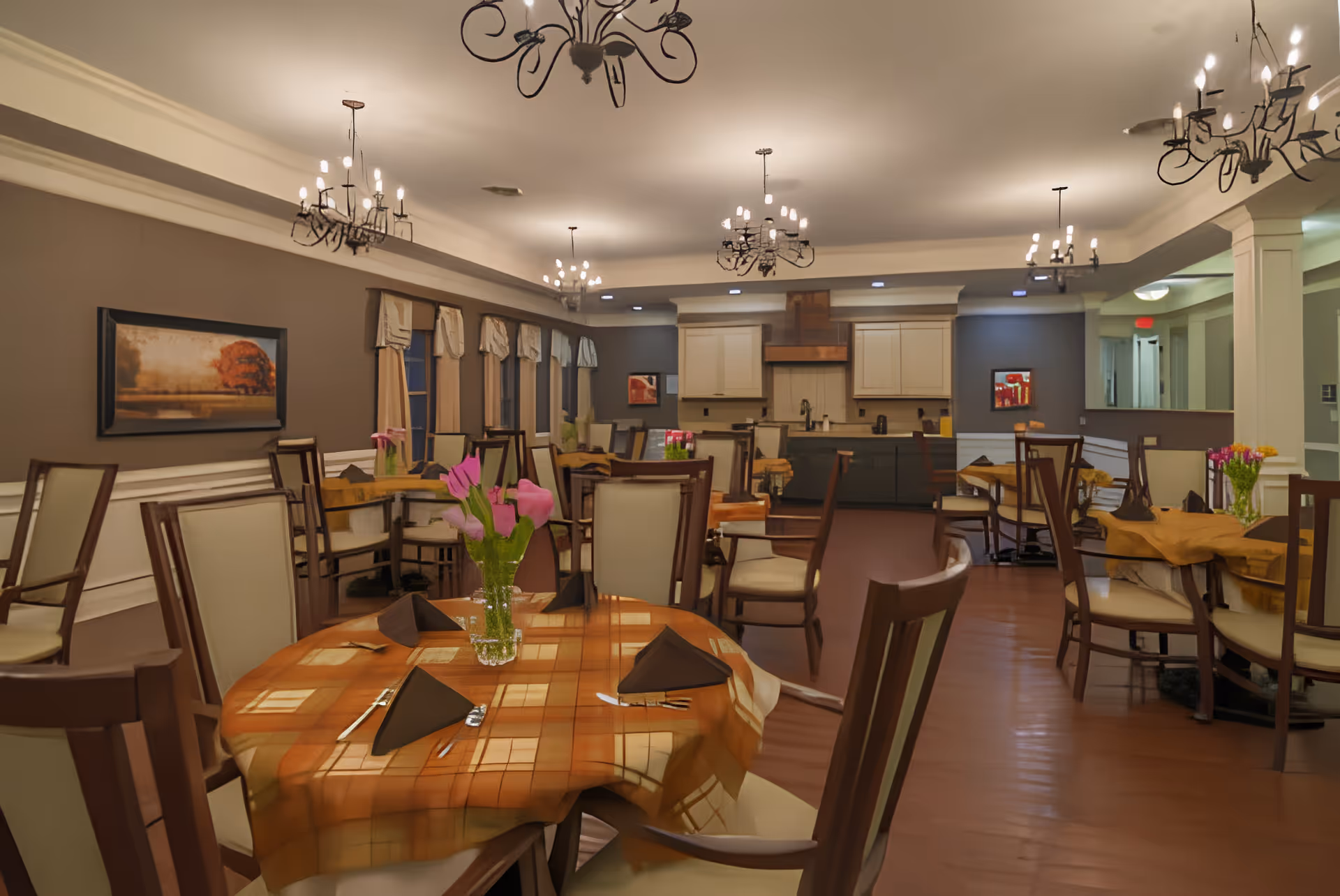 Well-lit dining room with multiple set tables, floral centerpieces, chandeliers, and a serving kitchen in the background.