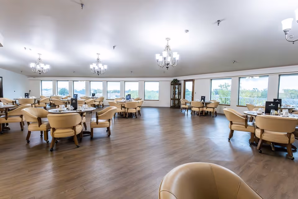 Spacious dining room with round tables and beige chairs on a wood floor under chandeliers and large windows.