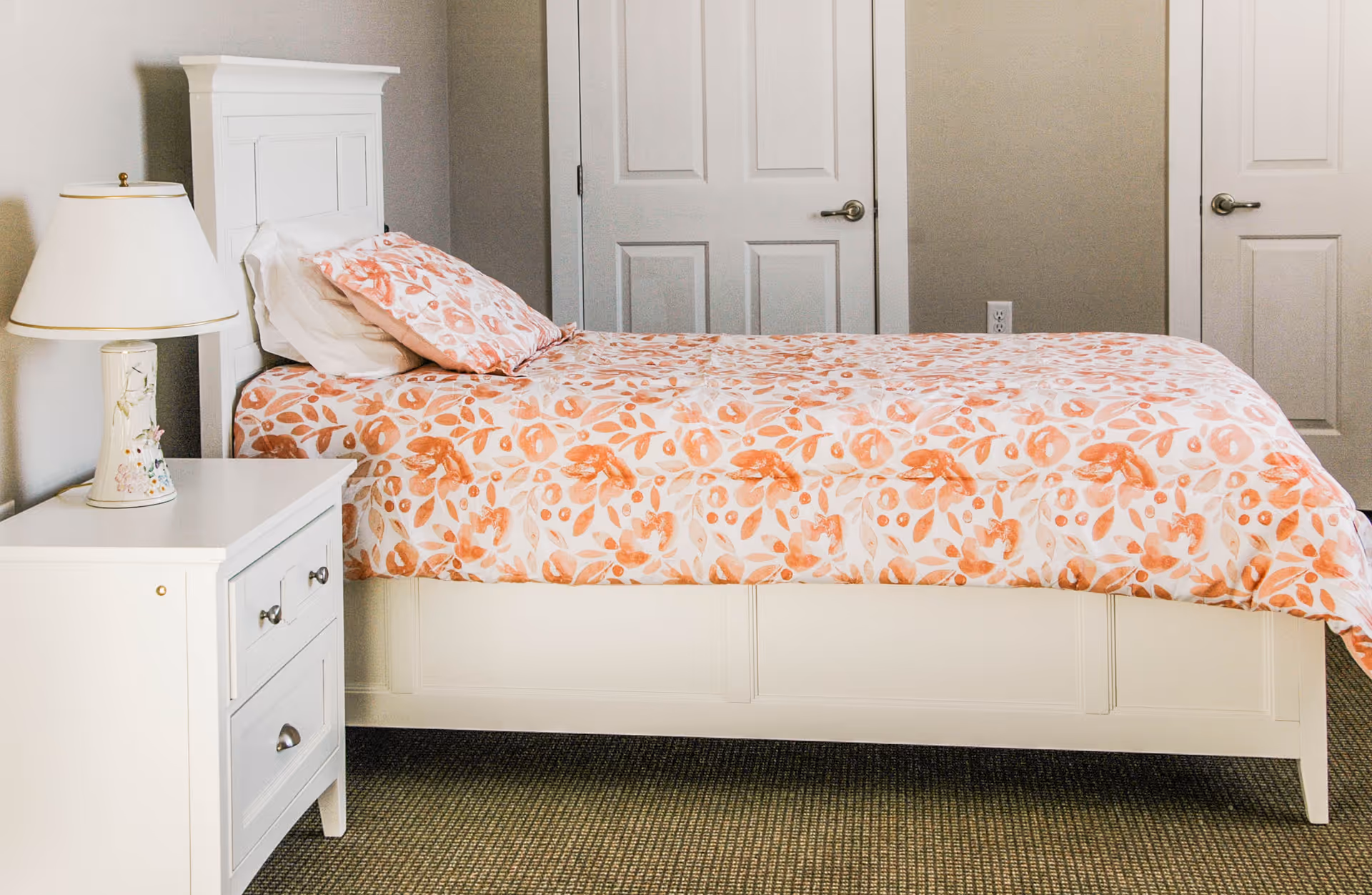 A neatly made single bed with a white wooden frame and a floral orange and white bedspread and pillow. Next to the bed is a white nightstand with two drawers and a decorative lamp on top. The background shows two closed white doors and a beige wall.