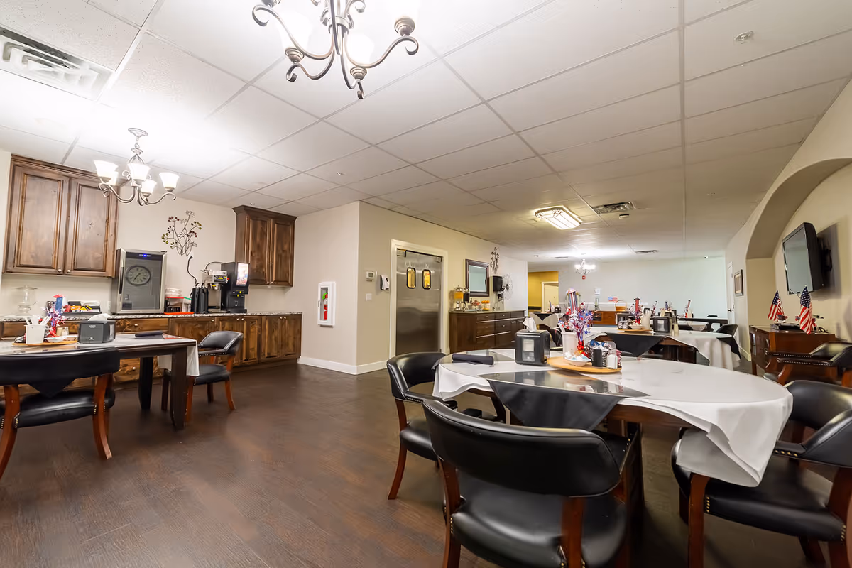 Interior view of a senior living facility dining room with several tables covered with white and black tablecloths, black cushioned chairs, wooden cabinets, a coffee machine, and patriotic decorations including small American flags. The room has a drop ceiling with chandeliers and a wall-mounted TV.