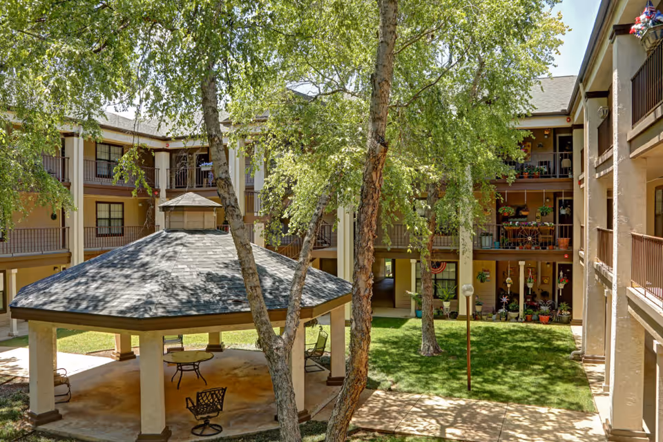 Outdoor courtyard area of a senior living facility with a large gazebo in the center, surrounded by trees and grass. The courtyard is enclosed by a three-story building with balconies decorated with plants and outdoor furniture.