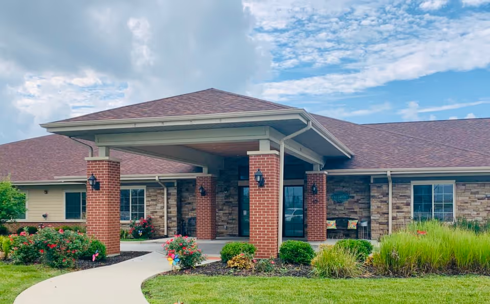 Front entrance of a single-story senior living facility with a covered porte-cochere, brick columns, and landscaped walkway.