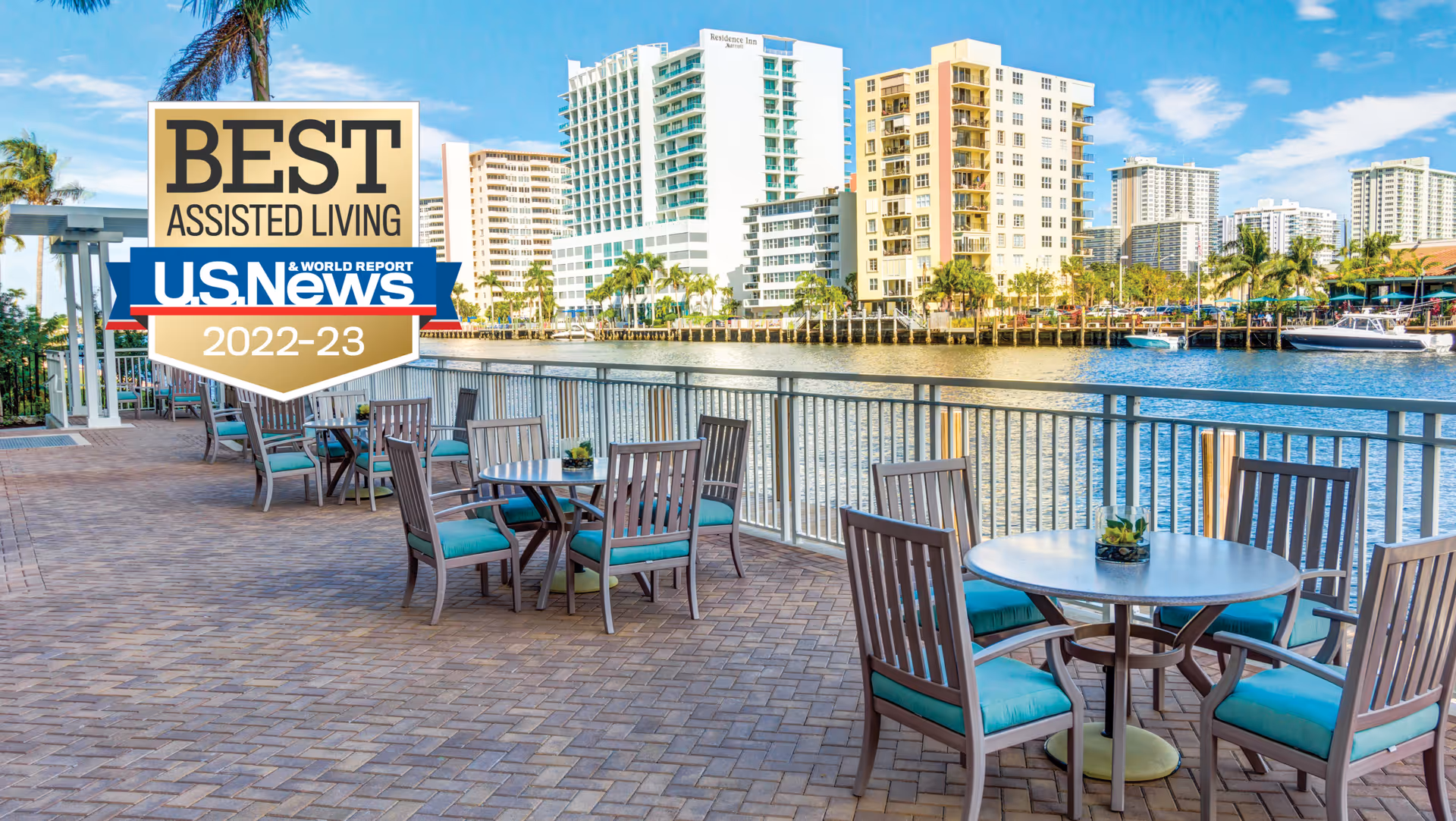 Outdoor patio area with several round tables and chairs with teal cushions overlooking a waterfront with boats and tall buildings in the background under a clear blue sky.