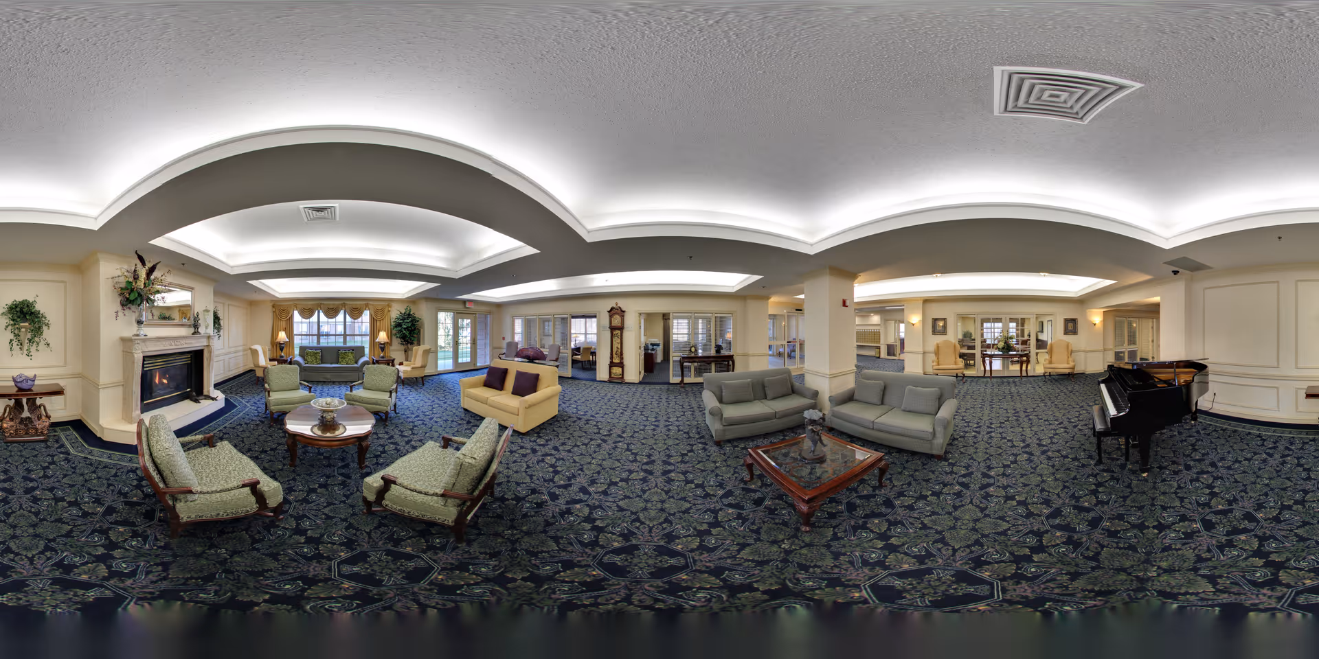 A spacious senior living community lounge with multiple seating areas including green upholstered chairs, beige sofas, and a glass coffee table. The room features a fireplace with decorative plants, a grand piano, and large windows allowing natural light. The ceiling has recessed lighting with a unique curved design, and the carpet has a dark floral pattern.