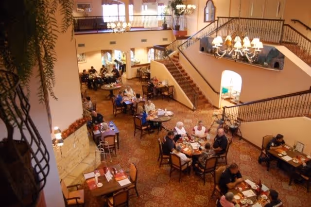 A spacious dining area in an assisted living facility with multiple tables where elderly residents are seated and enjoying their meals. The room features warm lighting, a carpeted floor, a staircase with wrought iron railings, and decorative plants. The atmosphere appears welcoming and comfortable.